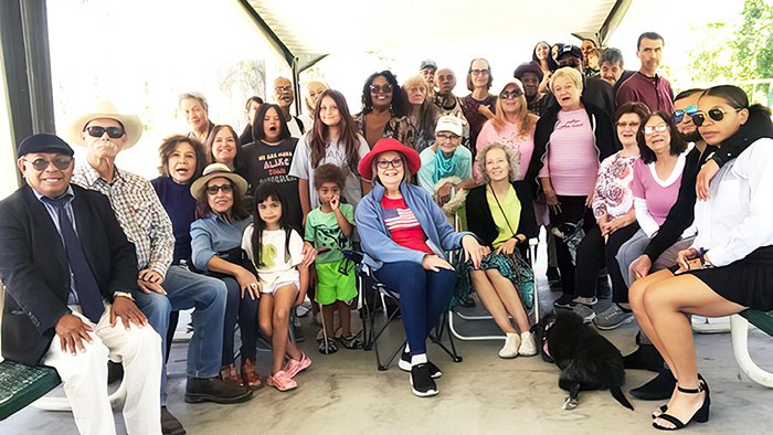 CHURCH COMMUNITY WEB A large, diverse group of people gathered under a covered outdoor pavilion for a social event. Individuals of various ages, from young children to older adults, are seated and standing in rows. Some sit on benches or folding chairs, and a few hold pets. The setting appears to be a park with trees in the background. Attendees wear casual clothing and many smile or look toward the camera, conveying a warm, communal atmosphere.