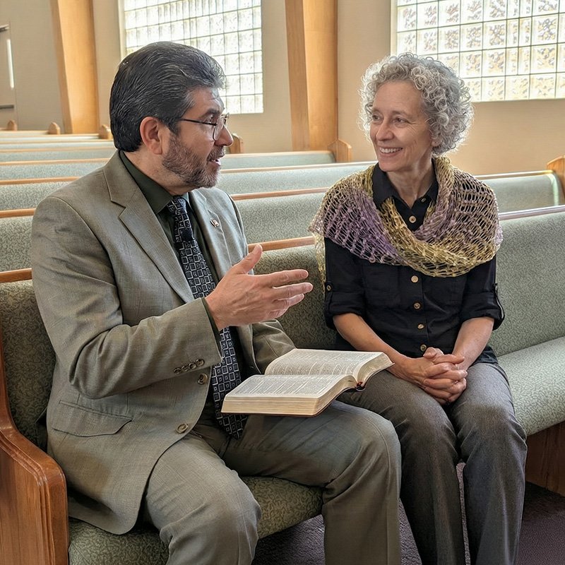 Two individuals sit in a church pew engaged in conversation. The person on the left wears a gray suit and tie, holds an open book—likely a Bible—and gestures with one hand. The person on the right, dressed in a dark shirt, gray pants, and a multicolored shawl, listens attentively. The setting includes wooden pews with green cushions and large glass block windows that let in natural light, suggesting a peaceful, reflective environment.