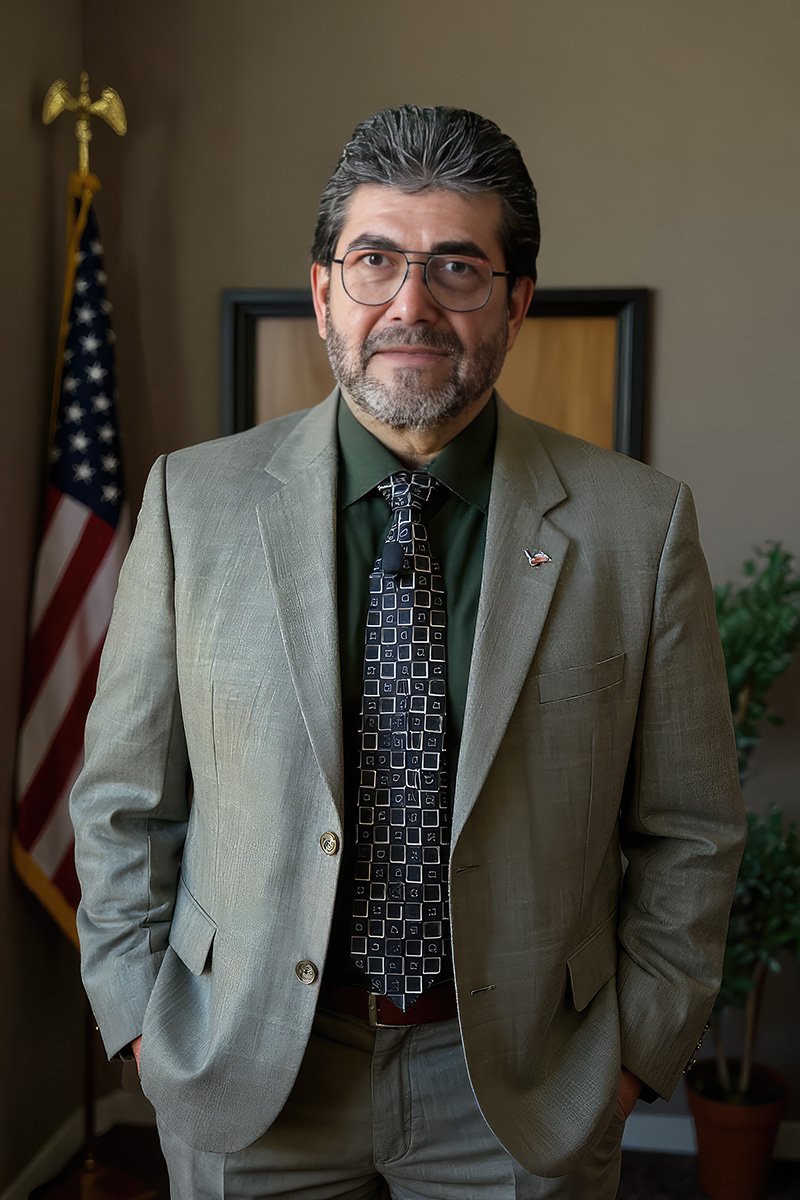 PASTOR SMILE v2 A professionally dressed individual stands indoors in front of a U.S. flag and a potted plant. They wear a light gray suit, a dark green shirt, and a patterned tie with square designs. A small lapel pin is visible on the left side of the suit jacket. The background includes a framed wooden panel and neutral-colored walls, suggesting a formal or office setting. The image conveys a poised, official presence, possibly in a governmental or administrative role.