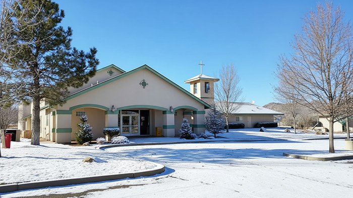 WINTER CHURCH Church building with light-colored walls and green trim, set in a snowy landscape under a clear blue sky. The entrance features double glass doors, and a bell tower with a cross rises to the right. Leafless trees and evergreens surround the property, evoking a peaceful winter atmosphere.