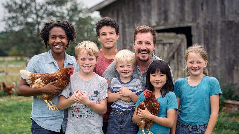 ADRA CHICKEN - Group Photo on Farm: A cheerful group of seven people—adults and children—stand in front of a rustic wooden barn. Three individuals hold chickens, suggesting a farm setting. The background features grass and a wooden fence, evoking a rural, community-oriented atmosphere.