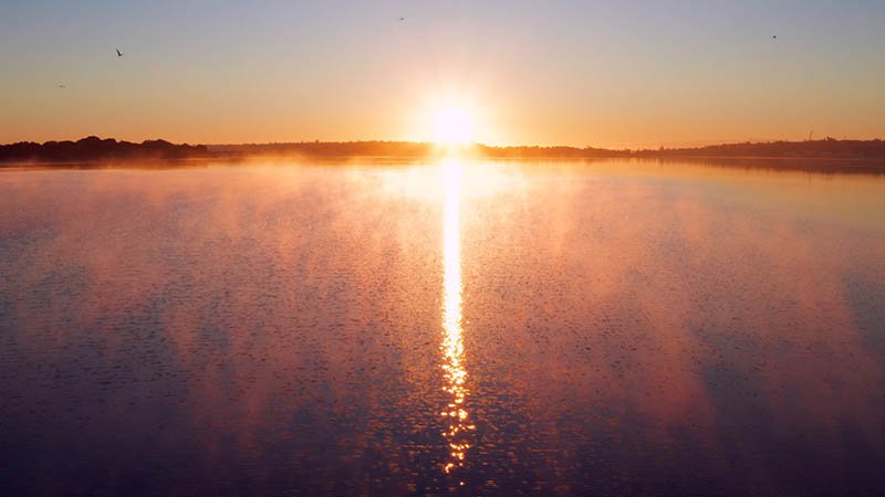 Serene sunrise over a misty body of water, with the sun low on the horizon casting a golden reflection across the calm surface. Light fog rises from the water, creating a soft, ethereal atmosphere. The sky transitions from deep blue to warm orange and yellow tones. A few birds fly overhead, and a distant shoreline is silhouetted against the morning light.