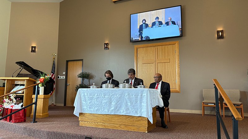 Interior of a church or religious meeting room during a communion service. Three formally dressed men sit behind a table covered with a white cloth, which holds silver communion trays. Behind them is a wooden wall with a carved cross and a large screen showing a live video feed of the same scene. To the left are a podium with a microphone, a piano, and an American flag. Floral arrangements decorate the podium and table. The room features beige walls, carpeted flooring, and wall-mounted lights