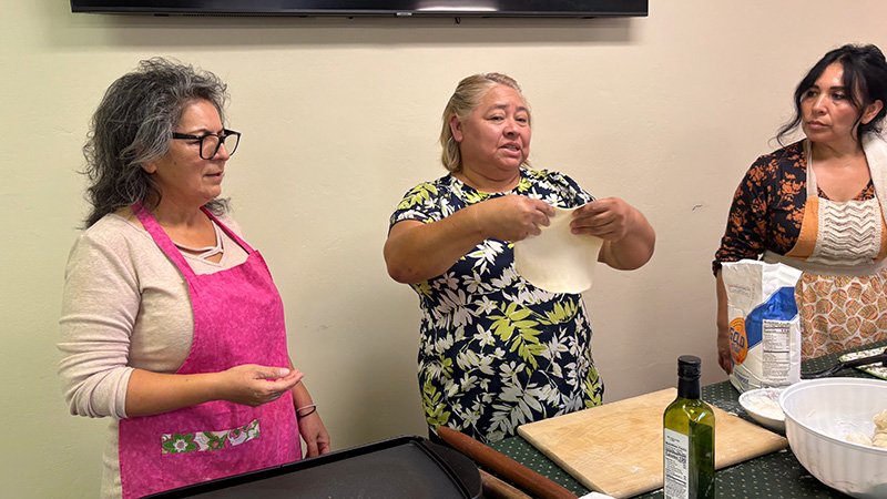Three women participating in a cooking activity indoors, possibly a class or demonstration. The woman in the center stretches a piece of dough, while the woman on the left, wearing a pink apron, observes. The woman on the right stands near a table with cooking supplies including olive oil, a rolling pin, a cutting board, a bowl of dough, and a flour bag. The setting resembles a community kitchen or classroom