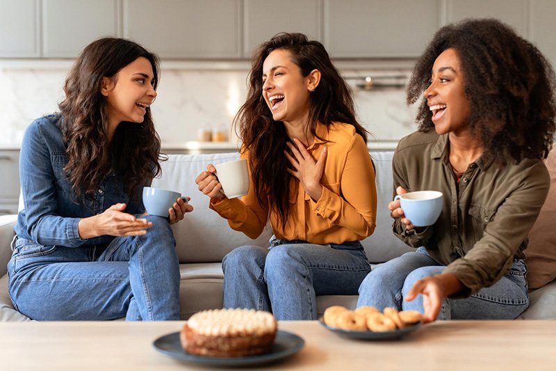 WOMEN HAVING TEA "Three diverse young women sitting on a couch, laughing and talking while holding mugs. A cake and a plate of cookies are on the coffee table in front of them in a bright, modern living room."