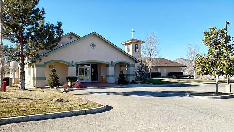 A beige church with green trim stands under a clear blue sky, featuring a bell tower topped with a cross. The building has arched entrances and is surrounded by neatly maintained trees and shrubs. A paved parking area sits in the foreground, with mountains visible in the background.