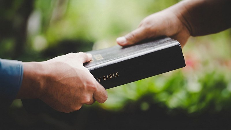 Two hands exchange a black Holy Bible with gold lettering against a blurred green outdoor background