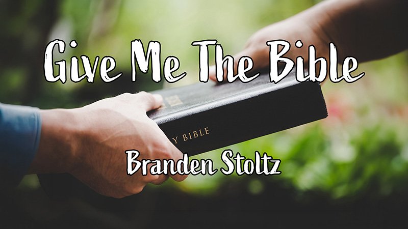 Two hands exchange a black Holy Bible with gold lettering against a blurred green outdoor background. White text over the image reads “Give Me The Bible” and below it “Branden Stoltz.”