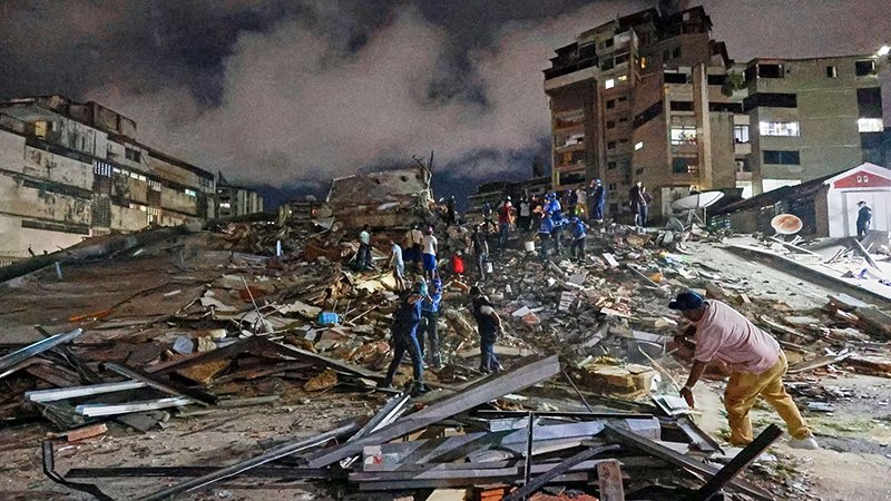 ADRA EARTHQUAKE A nighttime scene of a collapsed building with a large pile of debris. Several people are climbing over the rubble, searching through the wreckage under bright artificial lights. Surrounding structures show visible damage, and the atmosphere reflects an active rescue or recovery effort after a disaster.