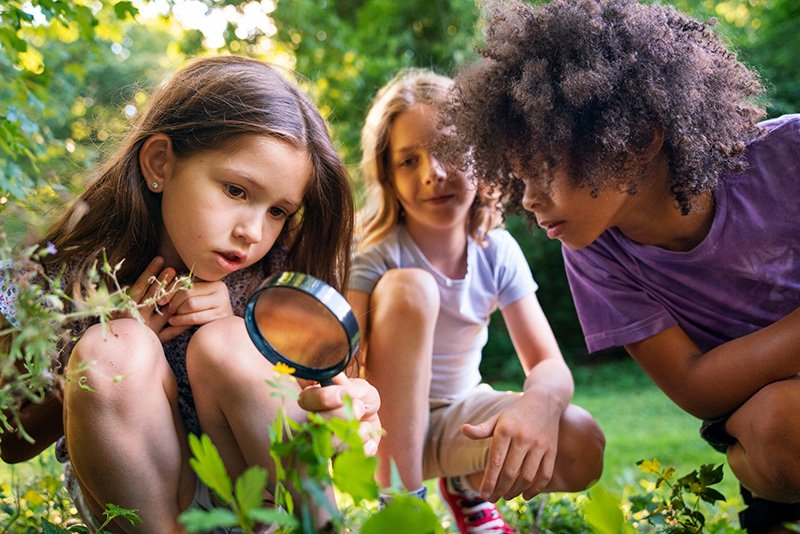 ADRA GARDENING Three children explore outdoors among green plants and trees. One child uses a magnifying glass to examine a plant closely while the other two watch with interest. The scene suggests a hands‑on nature or science activity.