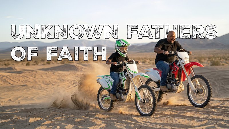UNKNOWS FATHERS Two people ride dirt bikes across a sandy desert, kicking up dust as they move. The rider on the left wears a helmet and rides a green and white bike, while the rider on the right rides a red and white bike without a helmet. Both are in casual clothing, with mountains visible in the distance under a clear sky. There is text behind the riders reading, "Unknown Fathers of Faith"